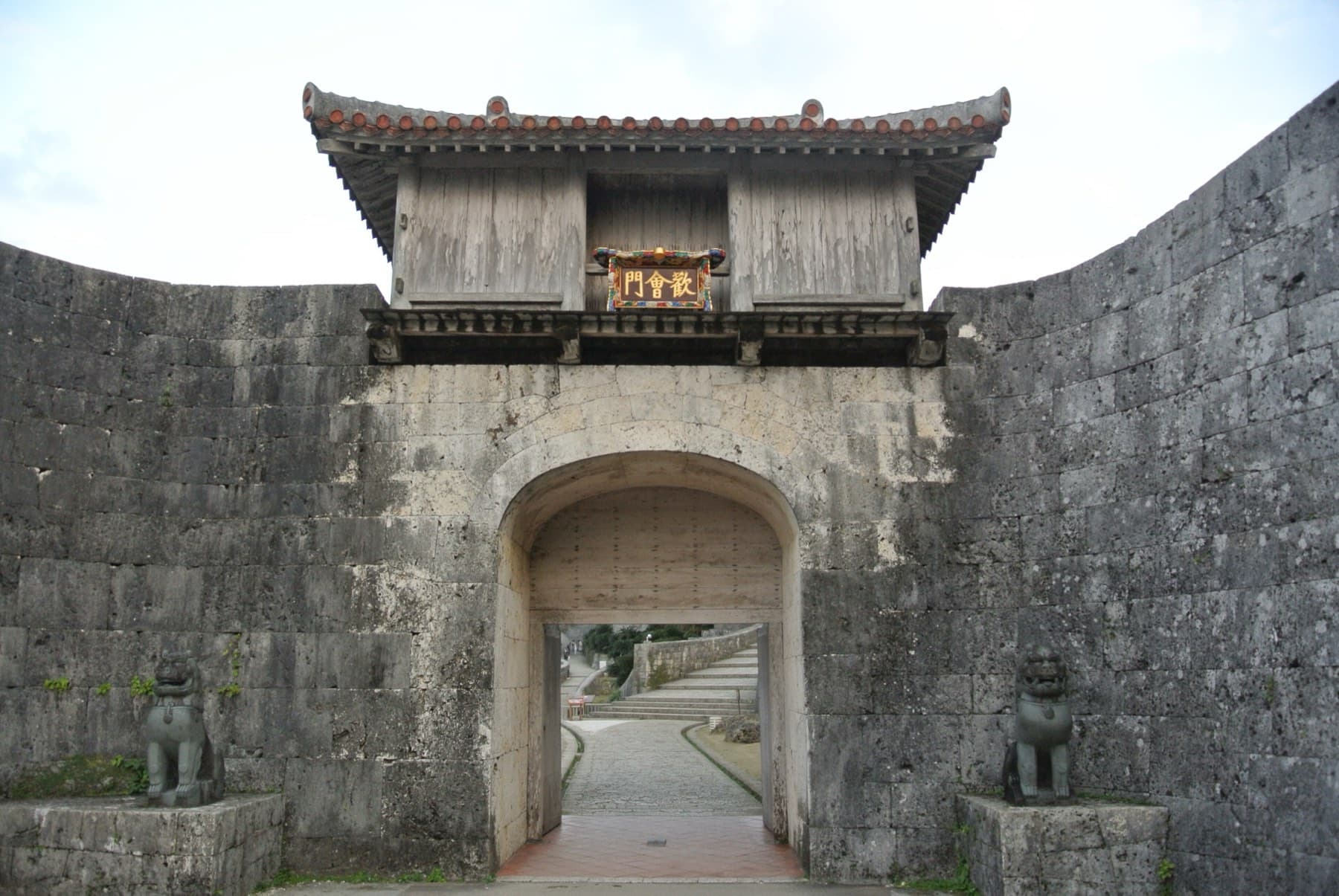 Two stone shisa guard the arched opening of Kankaimon gate, where a wooden tower sits on top of the curved limestone walls of Shuri Castle