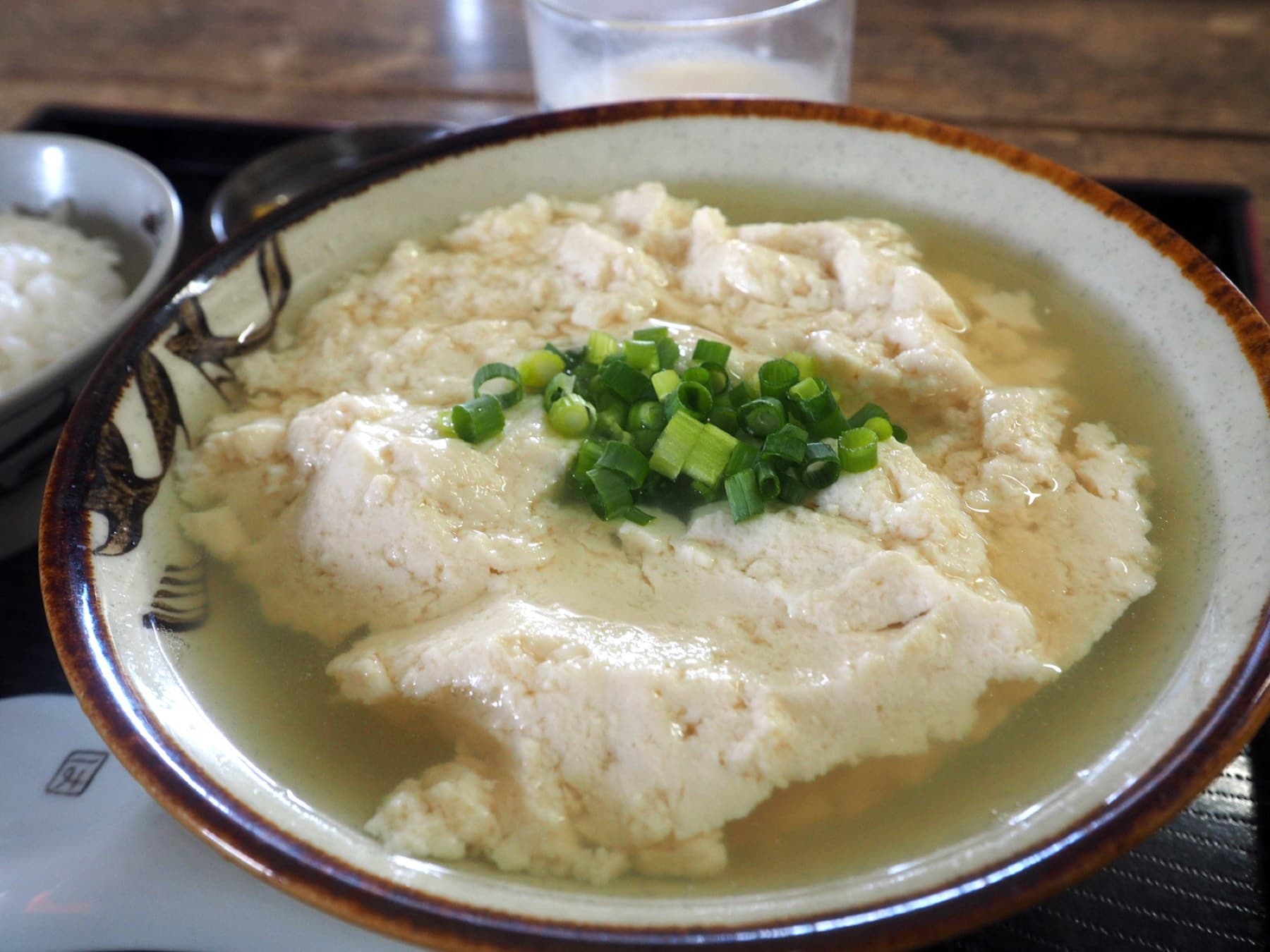 A wide bowl of yushi tofu at Tofu no Higa on Ishigaki holds loose white curds in pale broth with green onion, beside a small bowl of rice